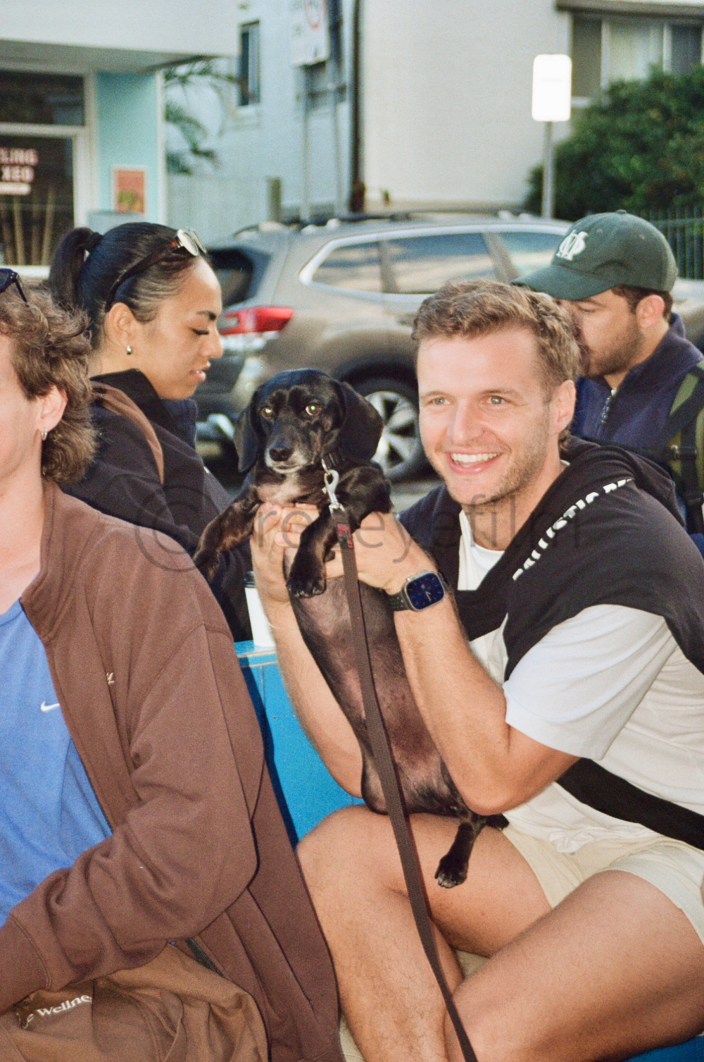 A group of people gathered outdoors, with one man smiling and holding a small black and tan dog, while others look on or are engaged in conversation.