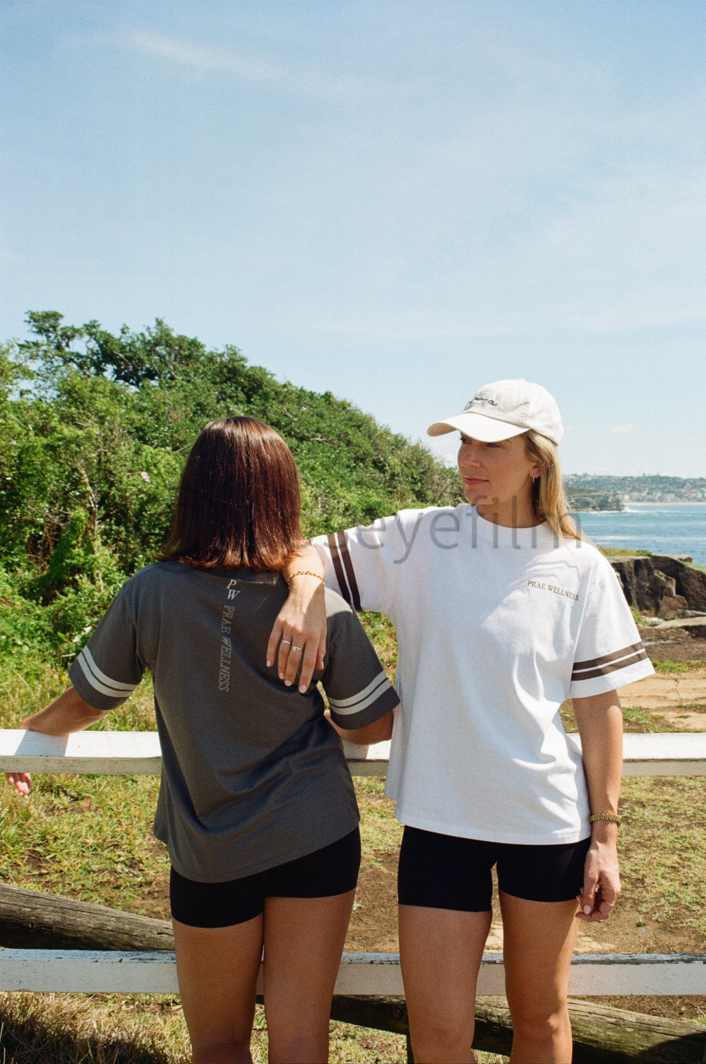 Two women standing outdoors near a white fence, with lush green trees and a body of water in the background, on a sunny day.