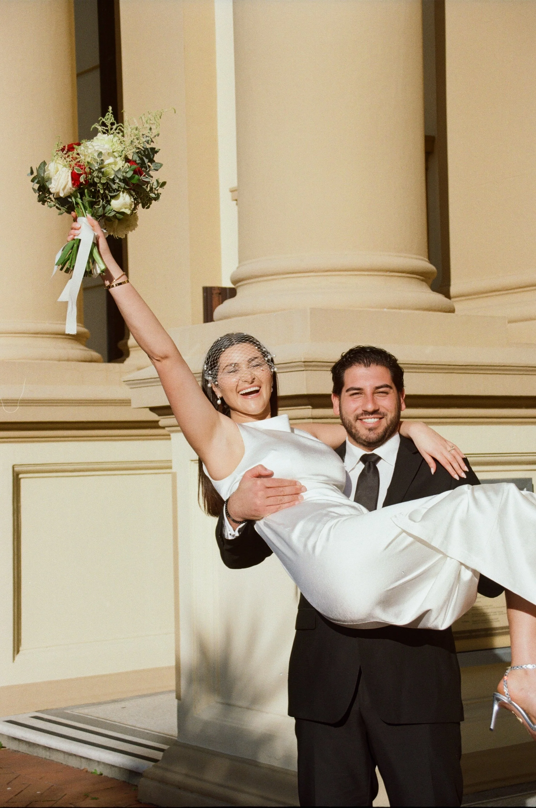 A smiling man in a black tuxedo lifts a woman in a white dress and veil, who is holding a bouquet of flowers, in front of a large beige column.