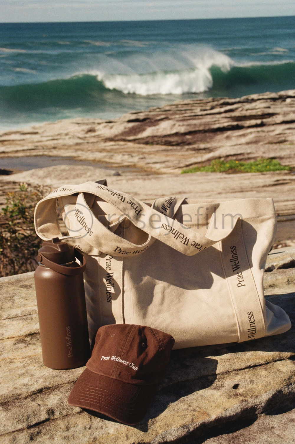 A beige tote bag, a brown water bottle, and a brown cap labeled 'Pure Wellness Club' on a rock near the ocean with waves in the background.