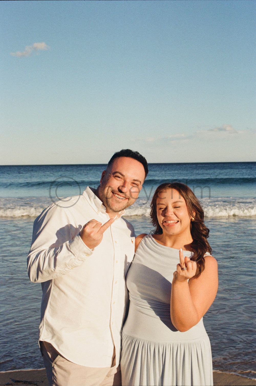 A man and woman on the beach with the ocean and sky behind them. They are smiling and making a rude gesture with their middle fingers.