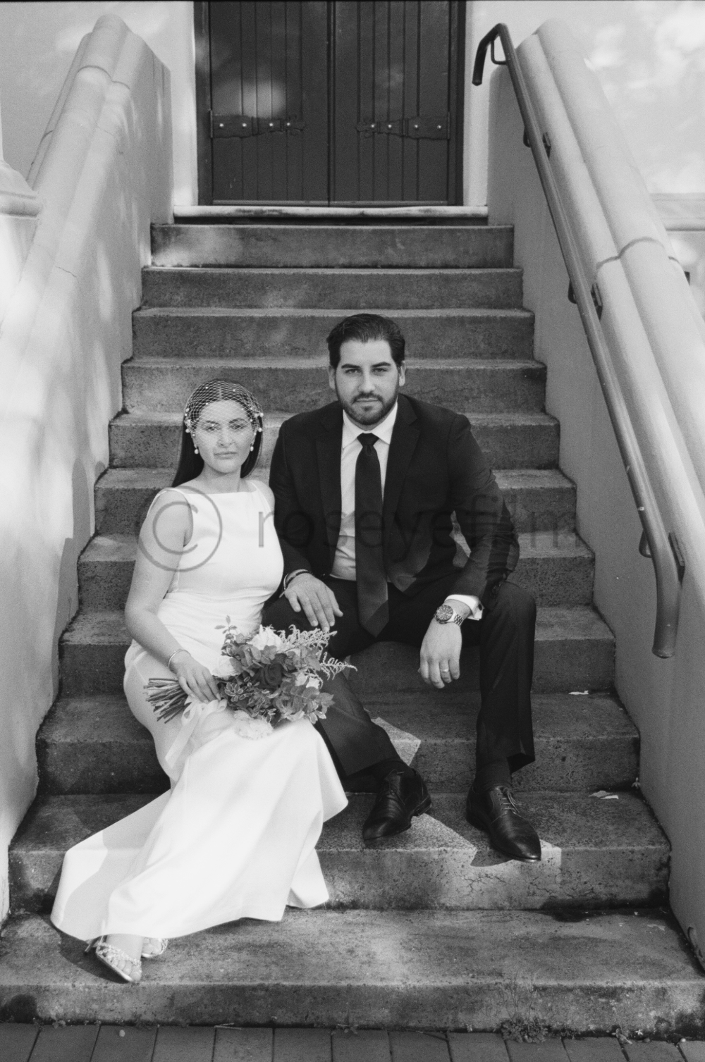 A bride and groom sitting on stairs outside the church, dressed in wedding attire, with the bride holding a bouquet and the groom wearing a watch and tie.