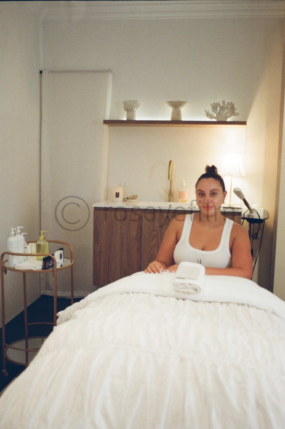 A woman sitting at a massage table in a spa room with minimal decor, a lamp, and a shelf with decorative items on the wall behind her.