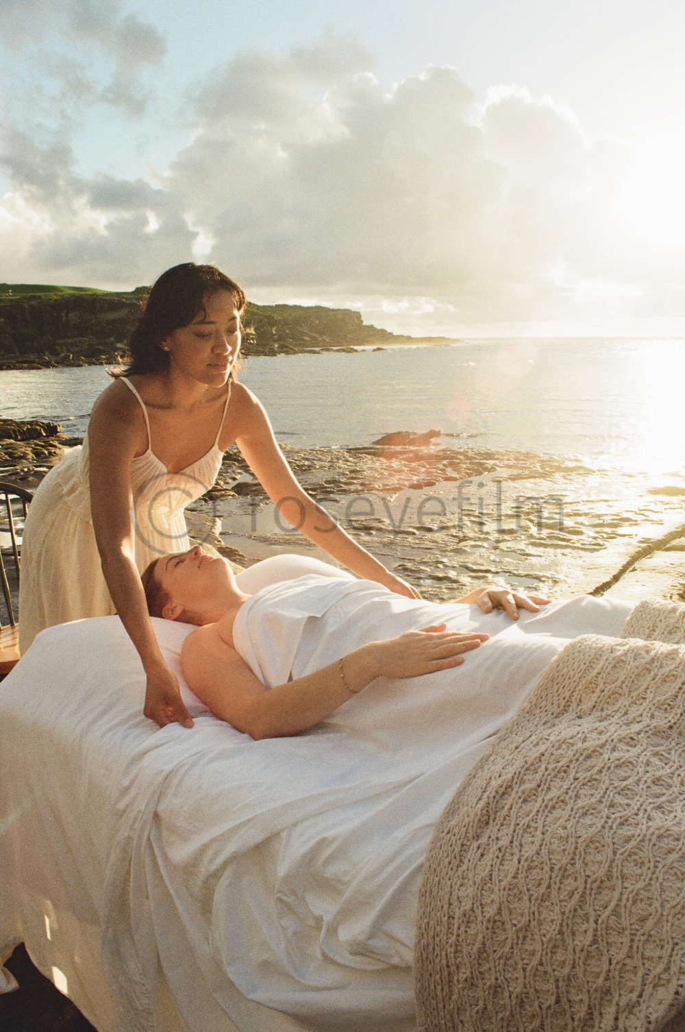 A woman lies in bed near the ocean while another woman tends to her, with the seaside and cloudy sky in the background.