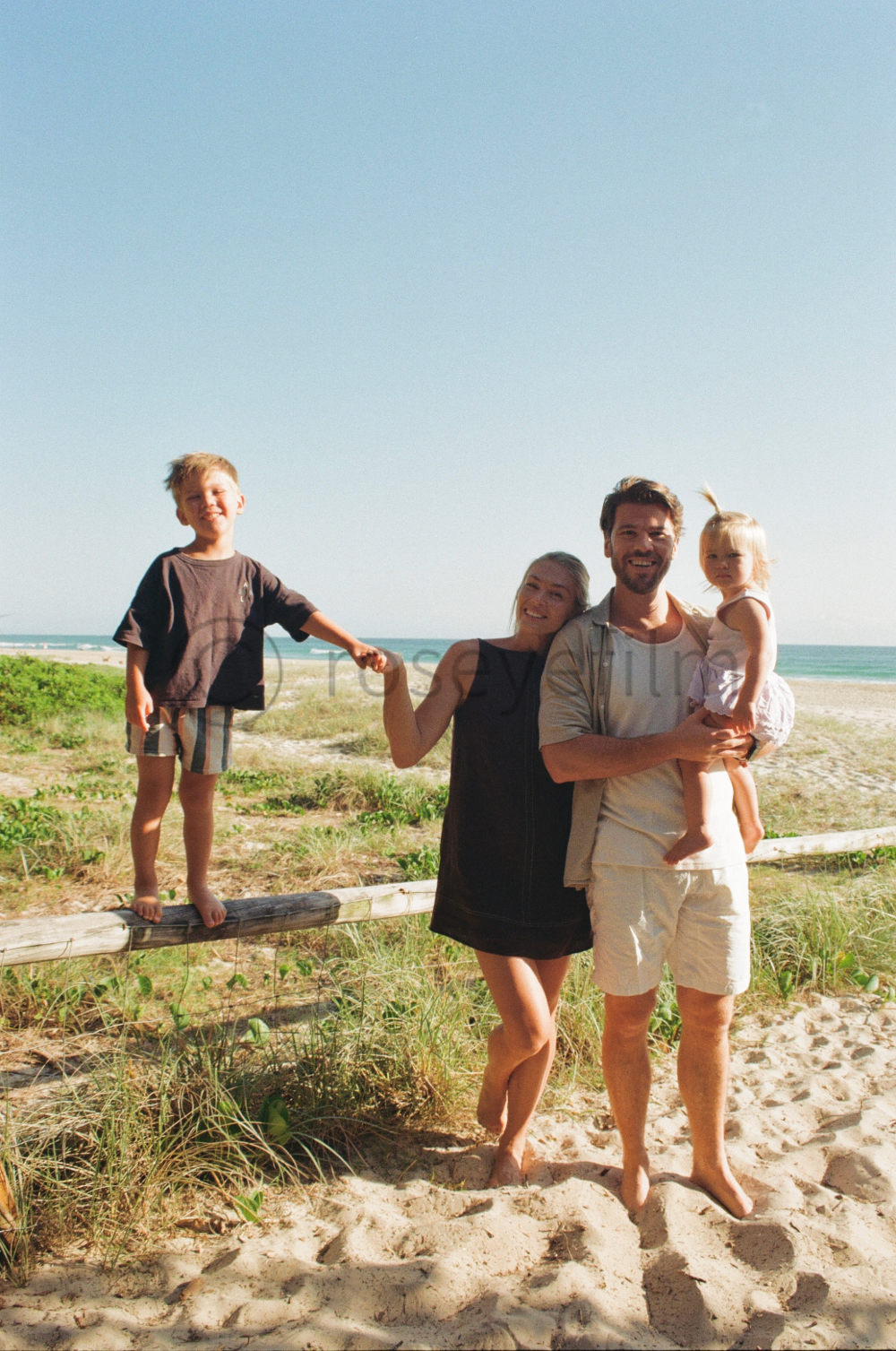 A family of four enjoying a day at the beach, standing on sandy ground with beach grass, the ocean, and a clear blue sky in the background. The young boy is standing on a wooden beam, the parents are holding their young daughter, smiling and relaxed.