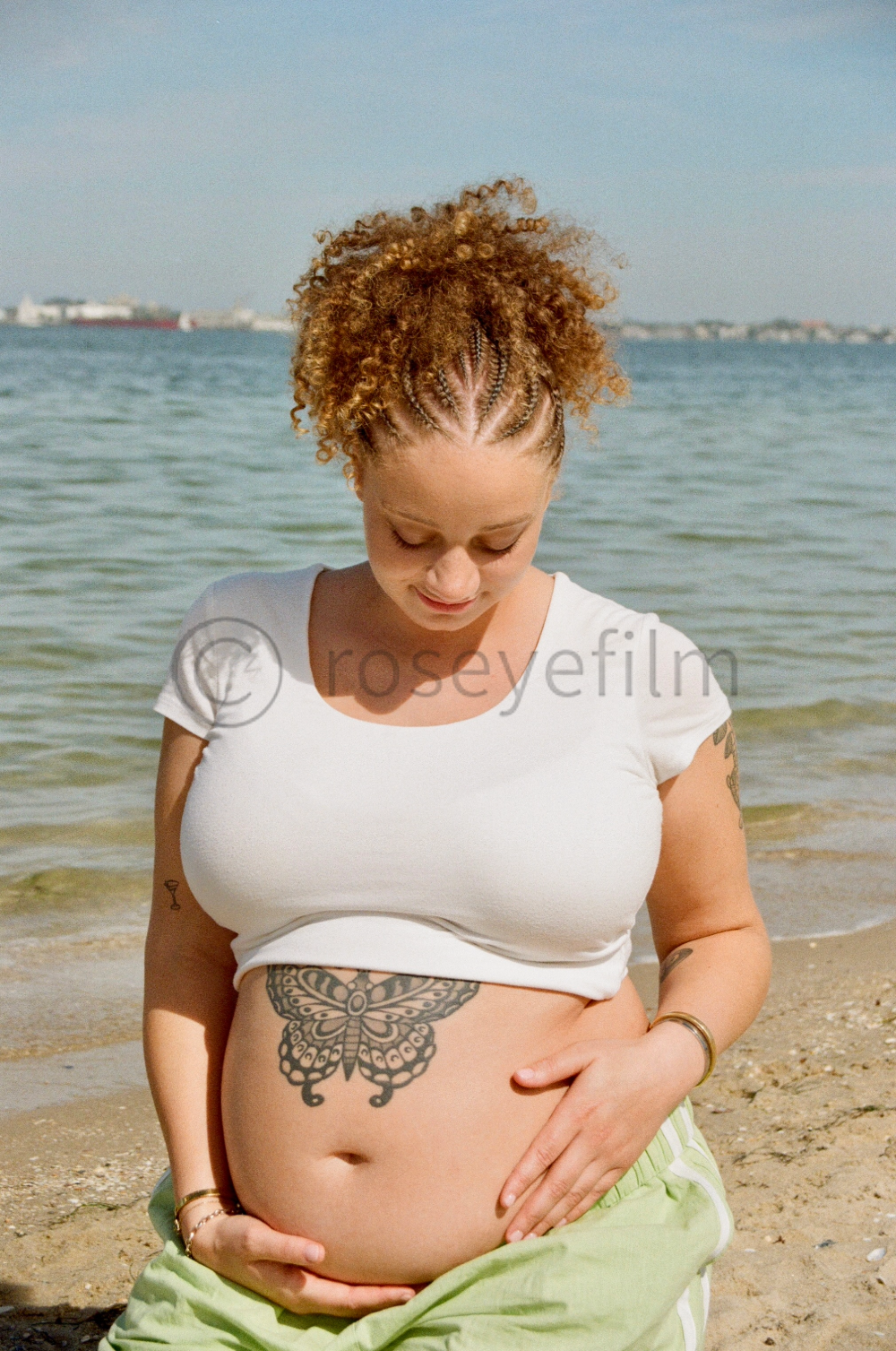 Pregnant woman with curly hair and tattoos on her arm and belly, standing on the beach looking down at her belly.