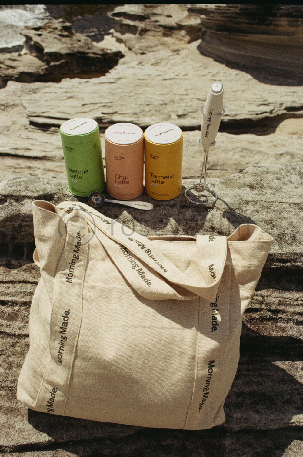 Travel essentials on a rock with a morning made tote bag, three cans of latte drinks (matcha, chai, turmeric), a white handheld whisk, and a small metal whisk attached to a stand, outdoors with rocky background.