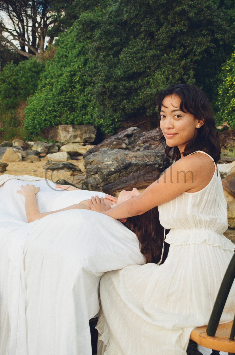 A woman in a white dress is sitting outdoors with another woman lying on a table, resting her head on the woman's lap. The scene is set near rocks and greenery, with trees and bushes in the background.