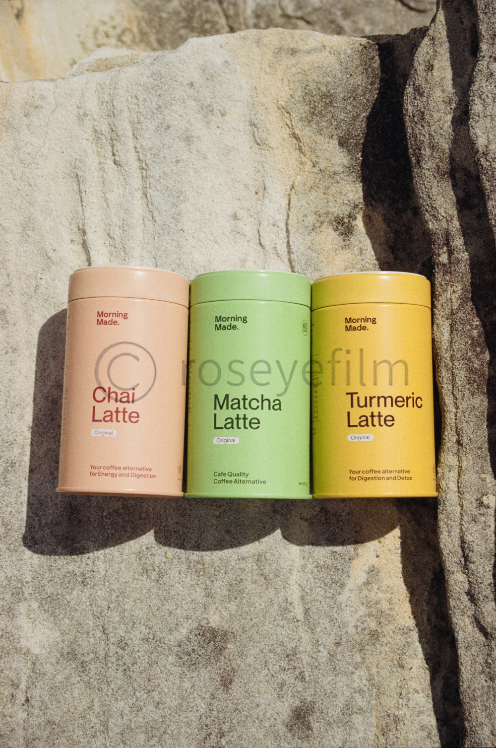 Three cans of herbal latte drinks in pastel colors, labeled Chai Latte, Matcha Latte, and Turmeric Latte, placed on a stone surface outdoors.