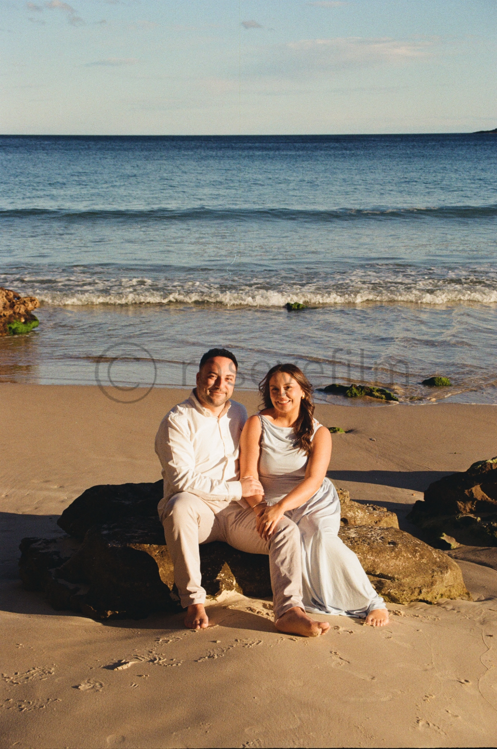 A couple sitting on rocks at the beach during sunset, with the ocean and sky in the background.