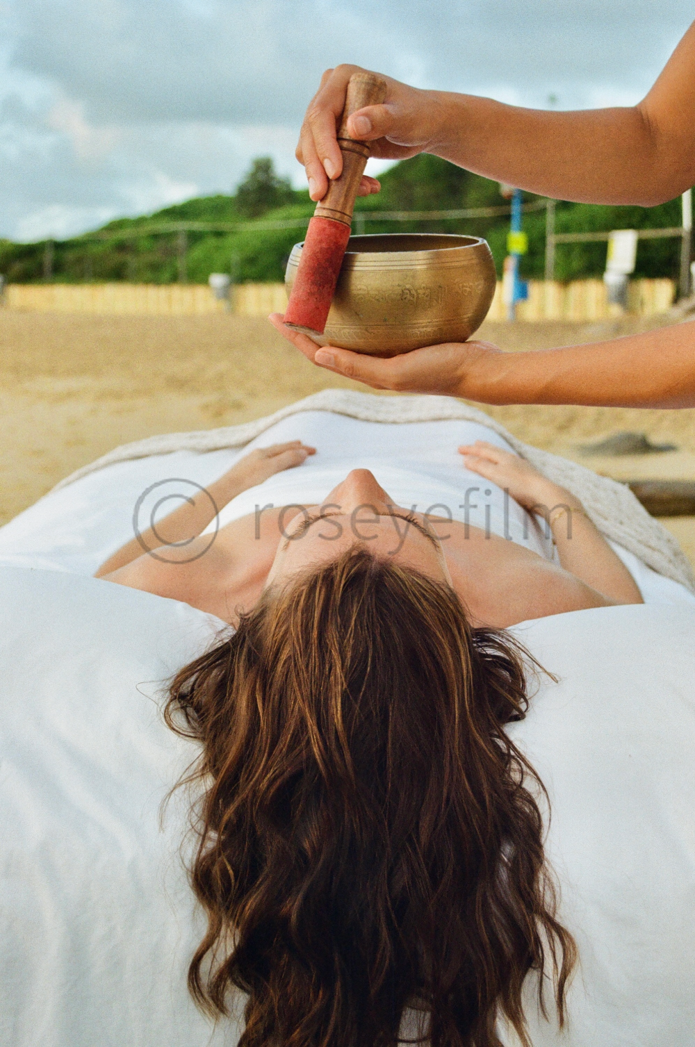 A woman lying on a massage table outdoors receiving a sound therapy with a Tibetan singing bowl and a small mallet.