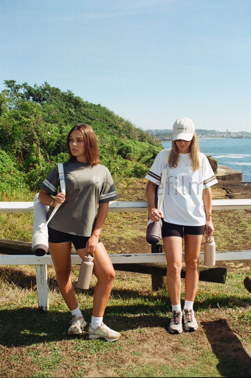 Two young women are standing outdoors near the coast with greenery and ocean in the background. They are dressed in athletic clothing with water bottles and yoga mats.