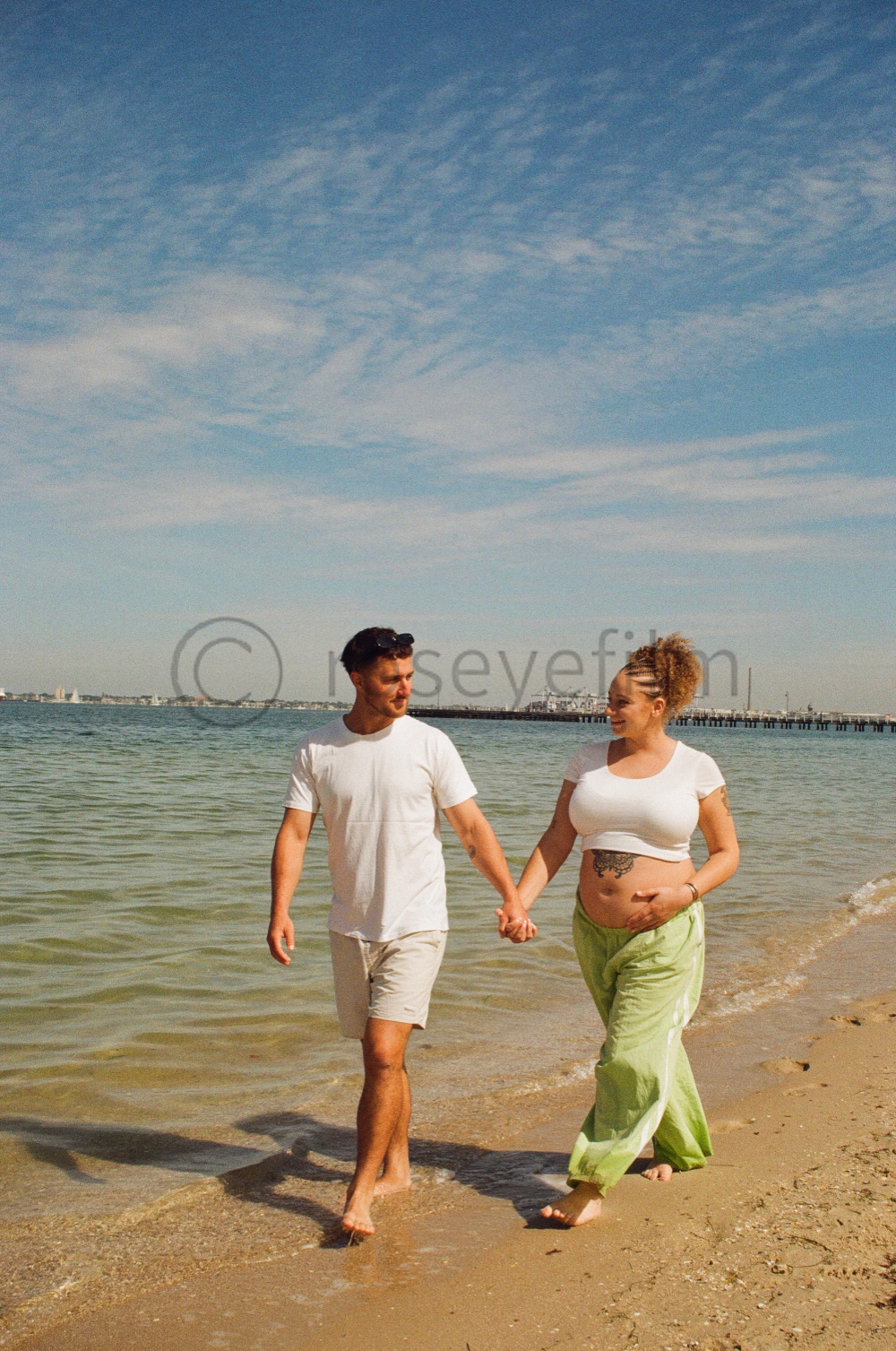 A couple walking hand in hand along the beach shore, with the woman visibly pregnant and smiling, beach, ocean, clear sky.