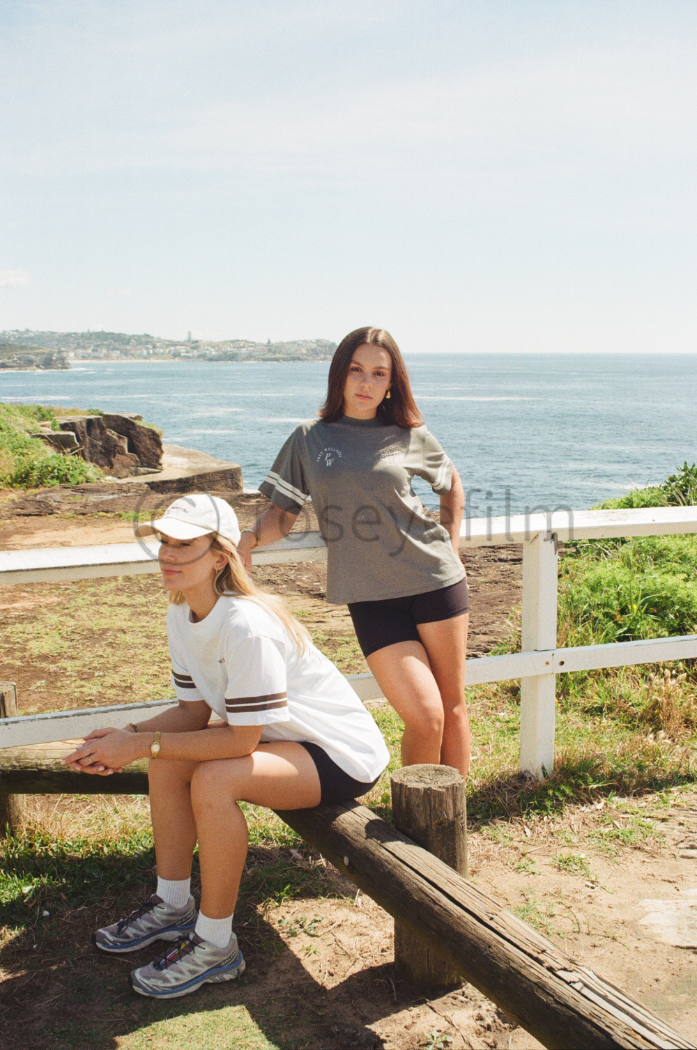 Two young women near a coastal area with ocean in the background. One woman is sitting on a wooden bench wearing a white baseball cap and white t-shirt, while the other is standing beside a white fence, wearing a grey t-shirt and black shorts.