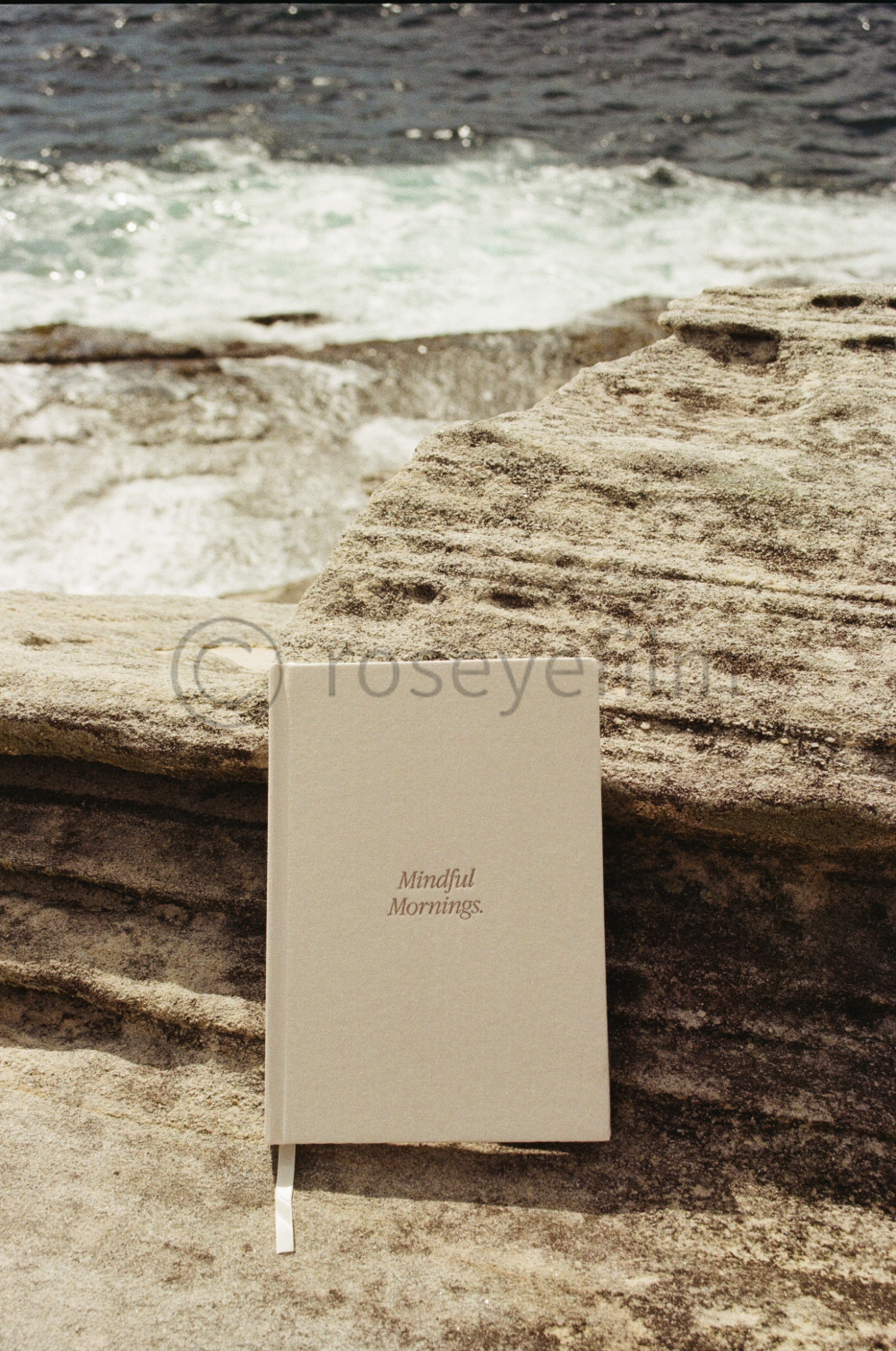 A beige notebook with the text 'Mindful Mornings.' resting against textured rocks on a beach, with waves and the ocean in the background.