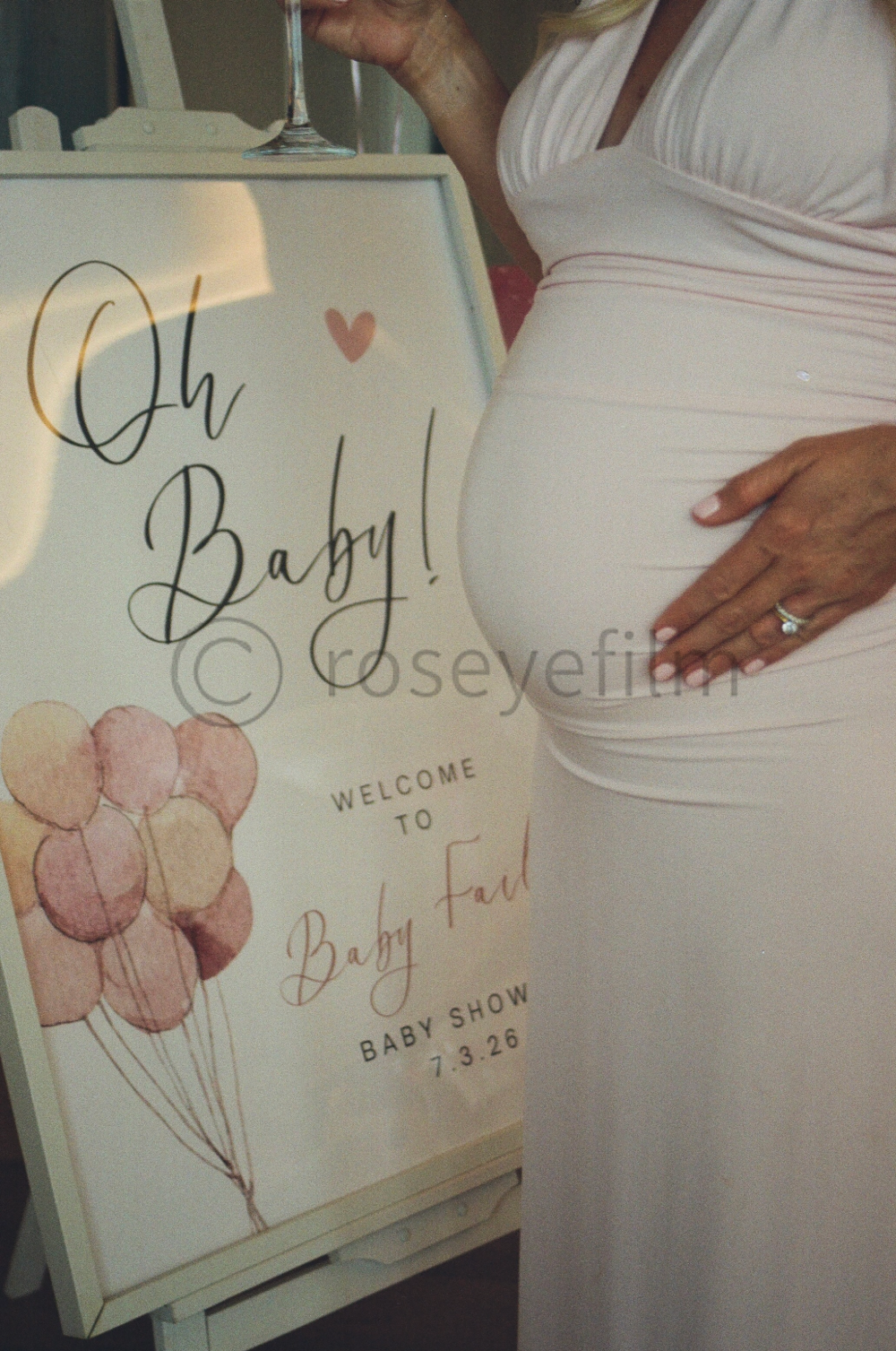 A pregnant woman in a white dress standing next to a decorative sign with the words "Oh Baby!" and illustrations of balloons, celebrating a baby shower event.