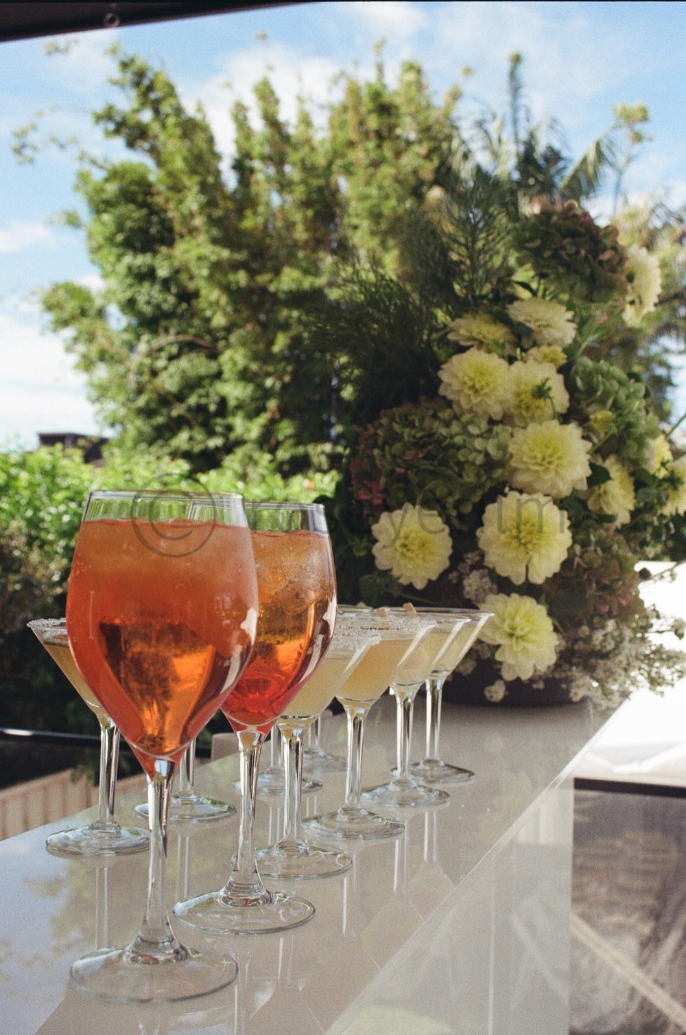 Set of glasses filled with pink and orange drinks on a white surface, with a floral arrangement and green trees and a blue sky in the background.