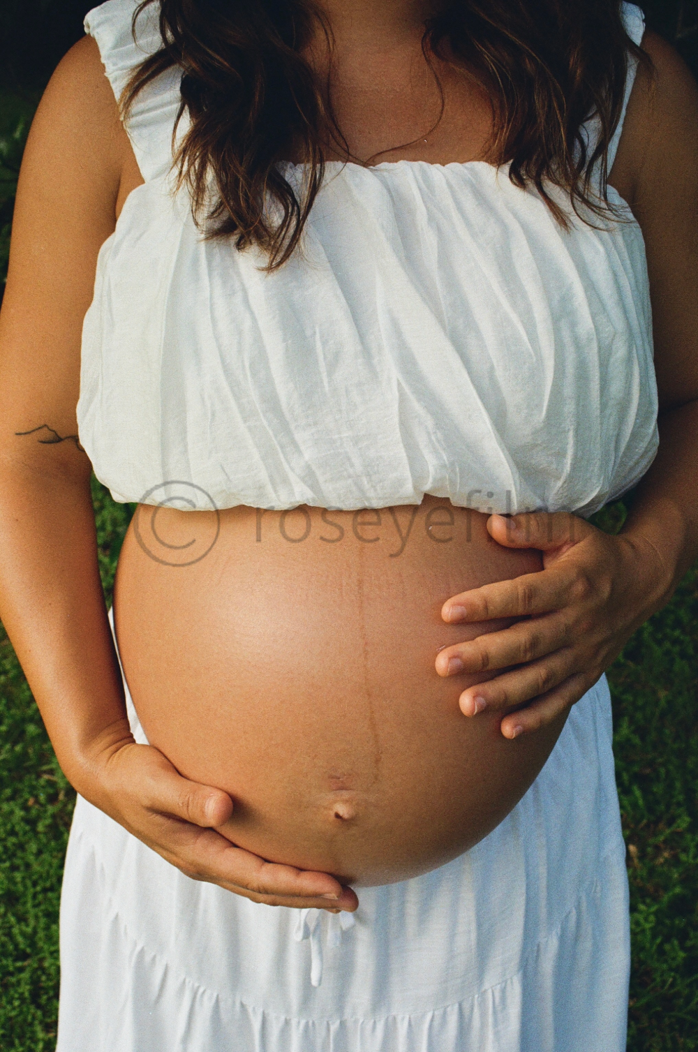 A pregnant woman holding her belly outdoors, wearing a white sleeveless top and a white skirt, with green grass in the background.