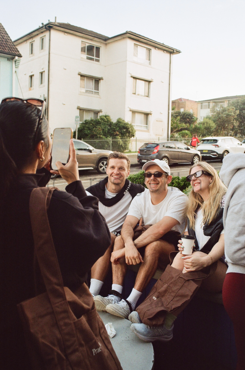 Group of four friends sitting outside, posing for a photo, with a woman taking their picture. They are smiling and appear to be enjoying a casual day.