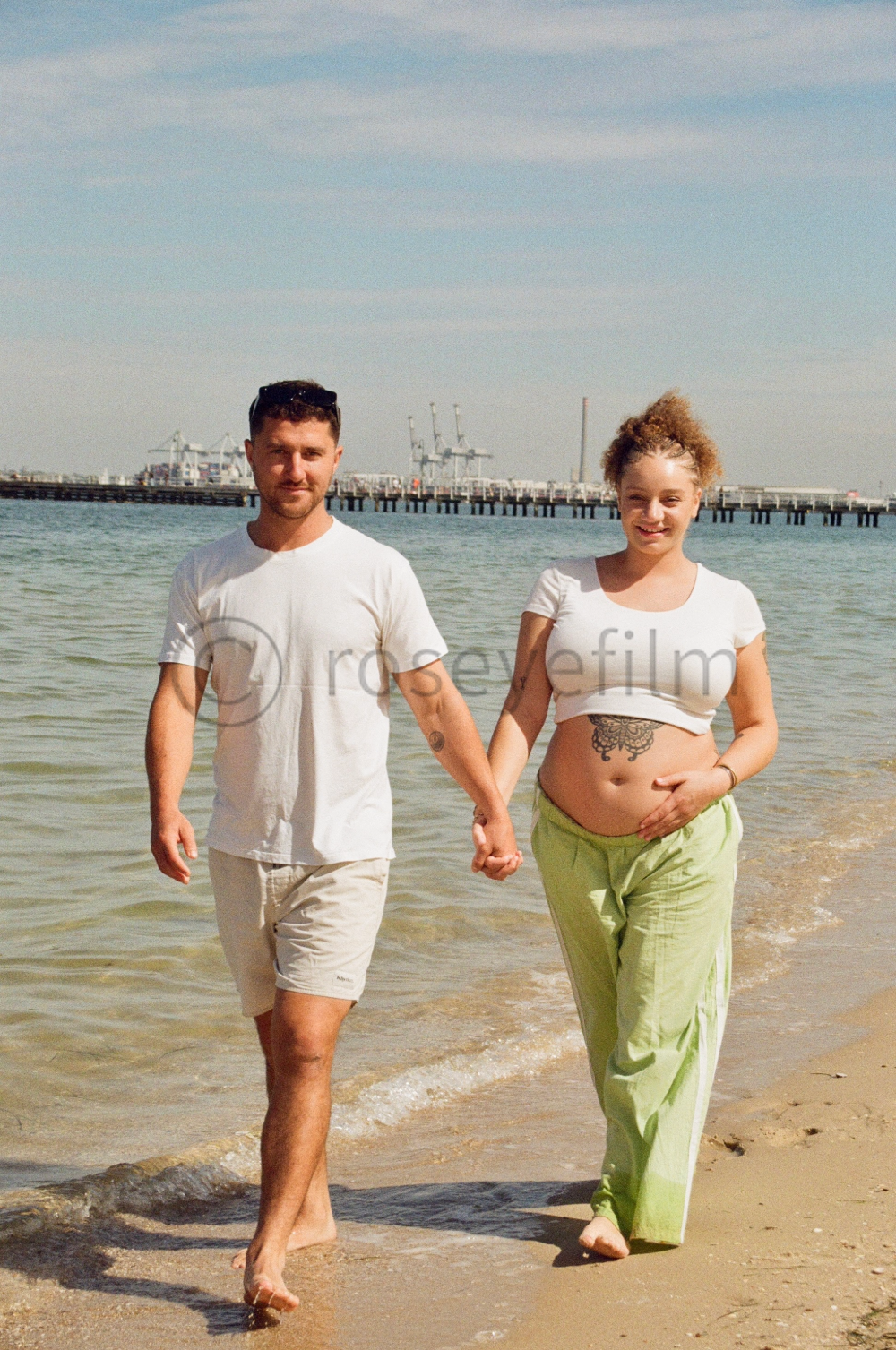 A couple walking hand in hand along the beach shoreline, near the water, with a pier and cranes in the background on a sunny day.