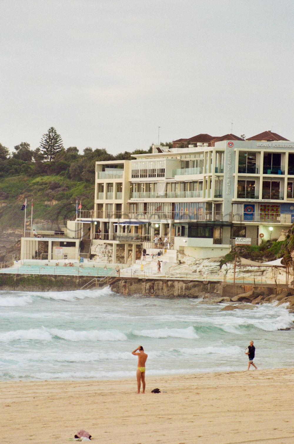 People on a beach, with a large modern building and hillside with trees in the background.