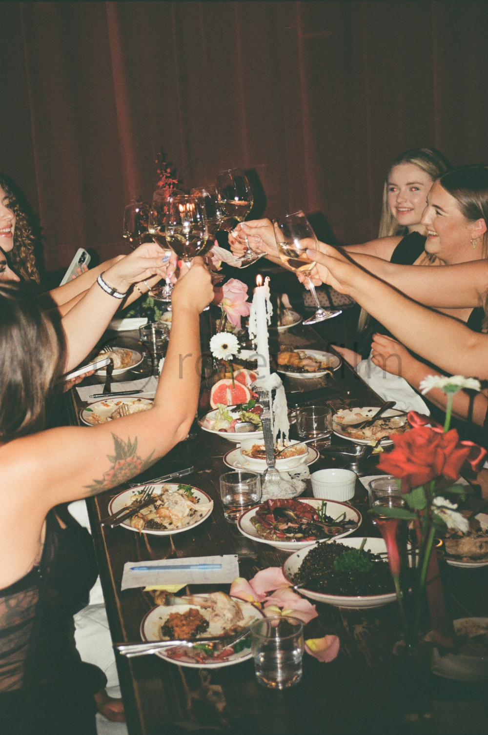 Group of women celebrating at a dinner table, raising glasses of white wine in a toast, with plates of food, floral decorations, and a lit candle in the center.