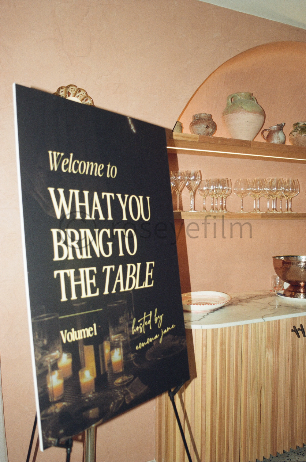 A welcome sign at a restaurant or event venue, with shelves holding ceramic vases and glassware in the background.
