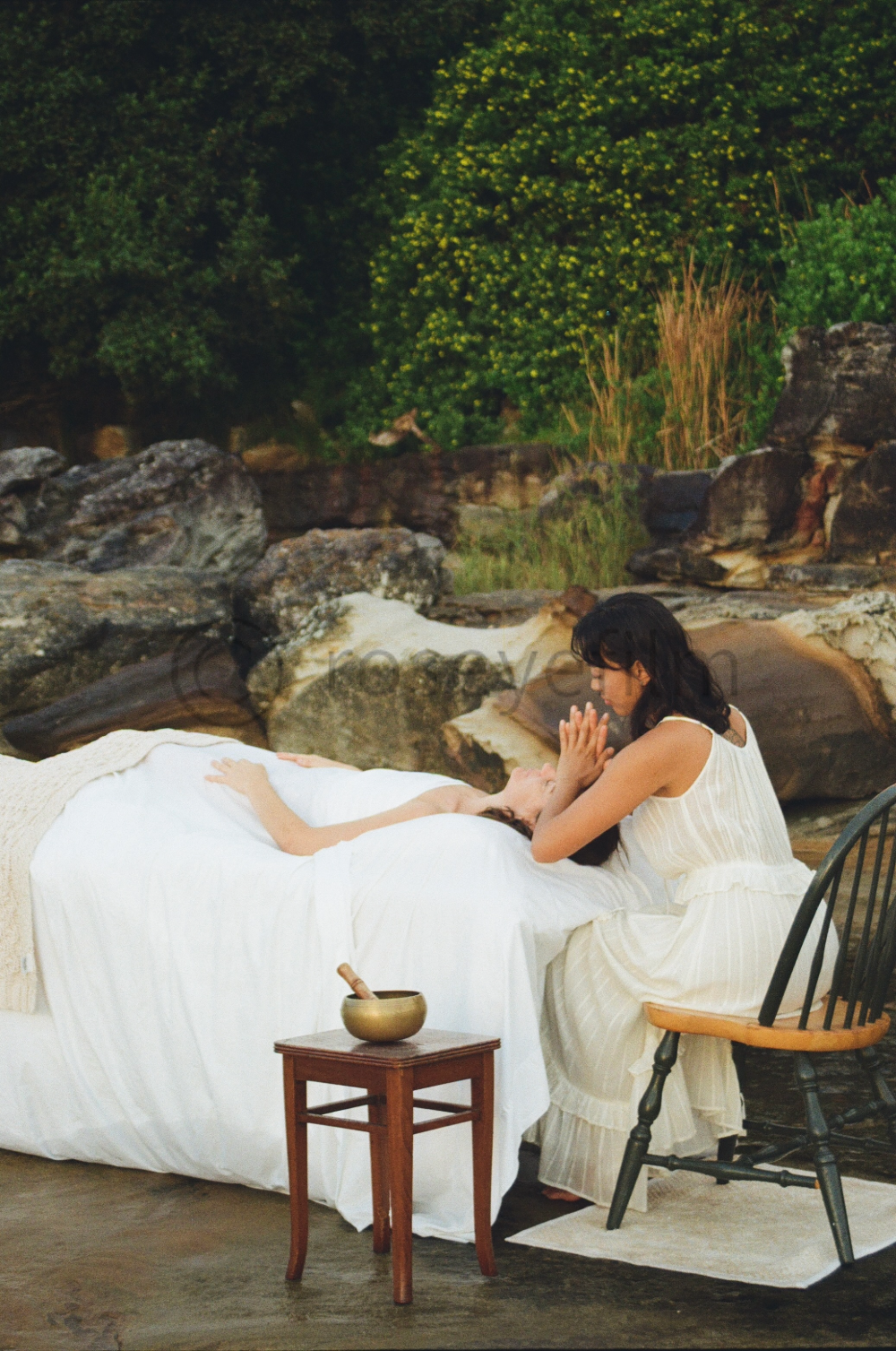 A woman in a white dress is kneeling beside another woman lying in bed outdoors, holding her hand and touching her face gently, with a backdrop of rocks and green bushes.