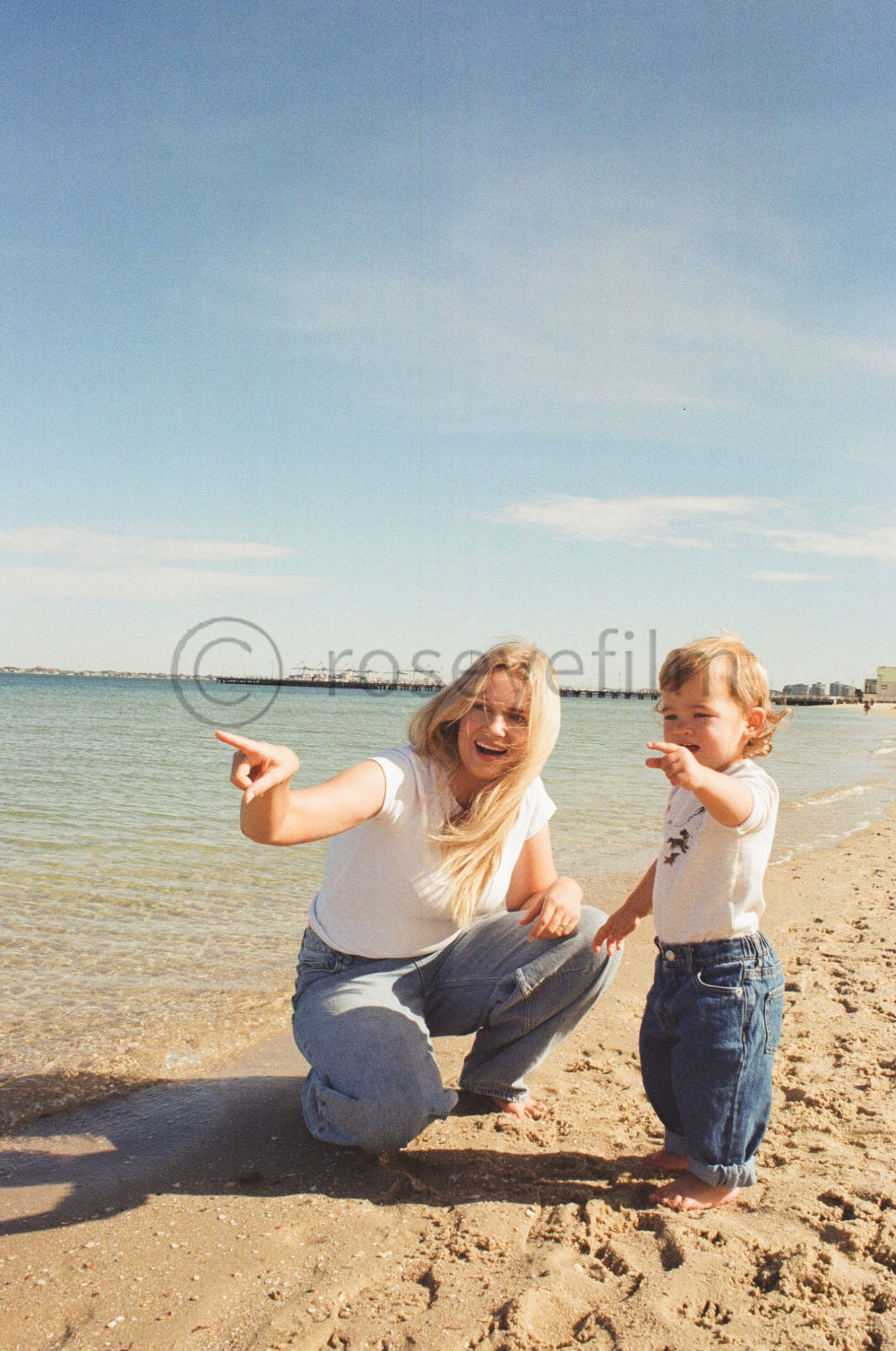 A woman and a young boy at the beach, both smiling and pointing.