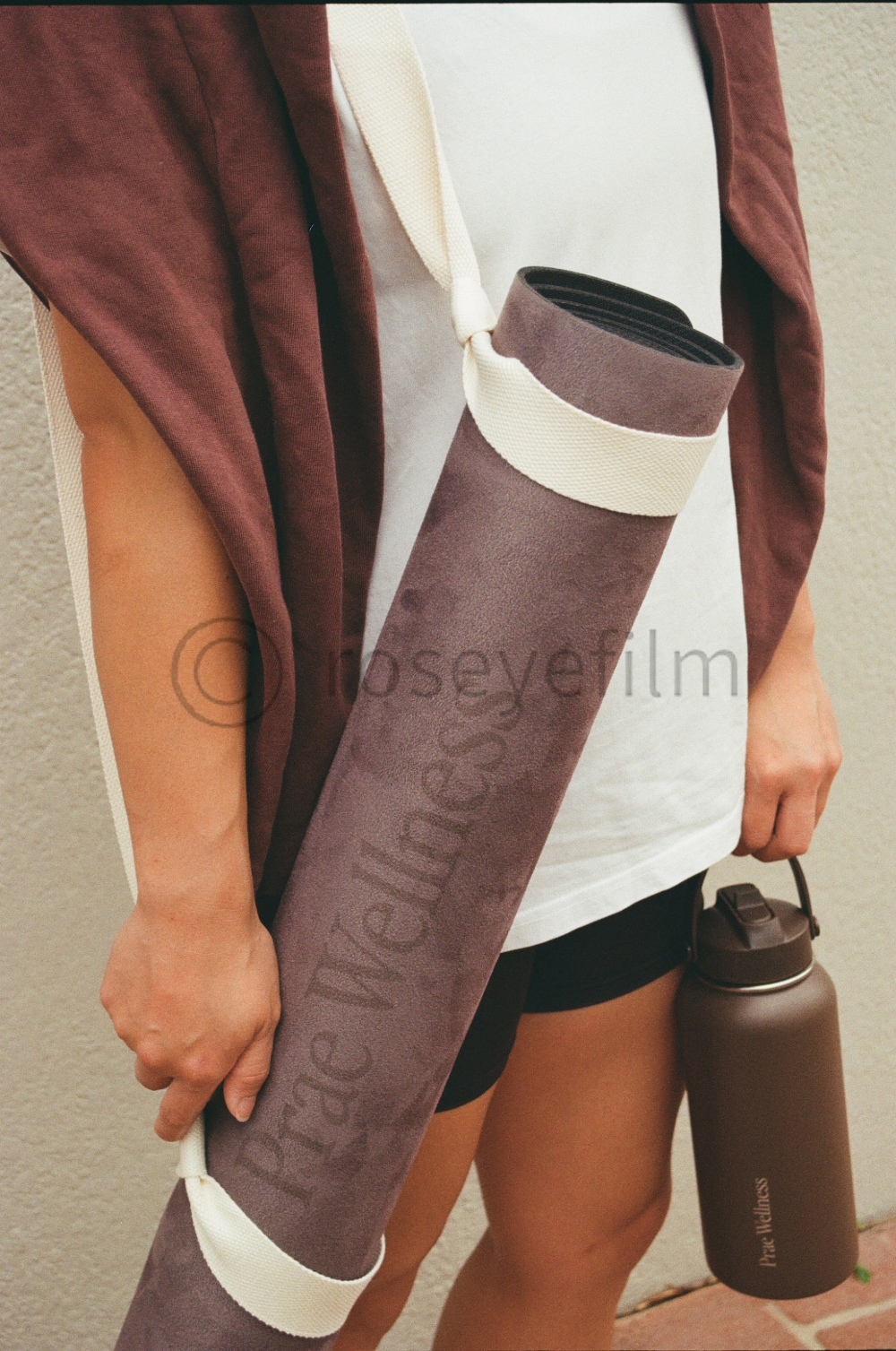 Person holding a yoga mat and water bottle, standing against a beige wall, wearing sports shorts and a loose tank top.