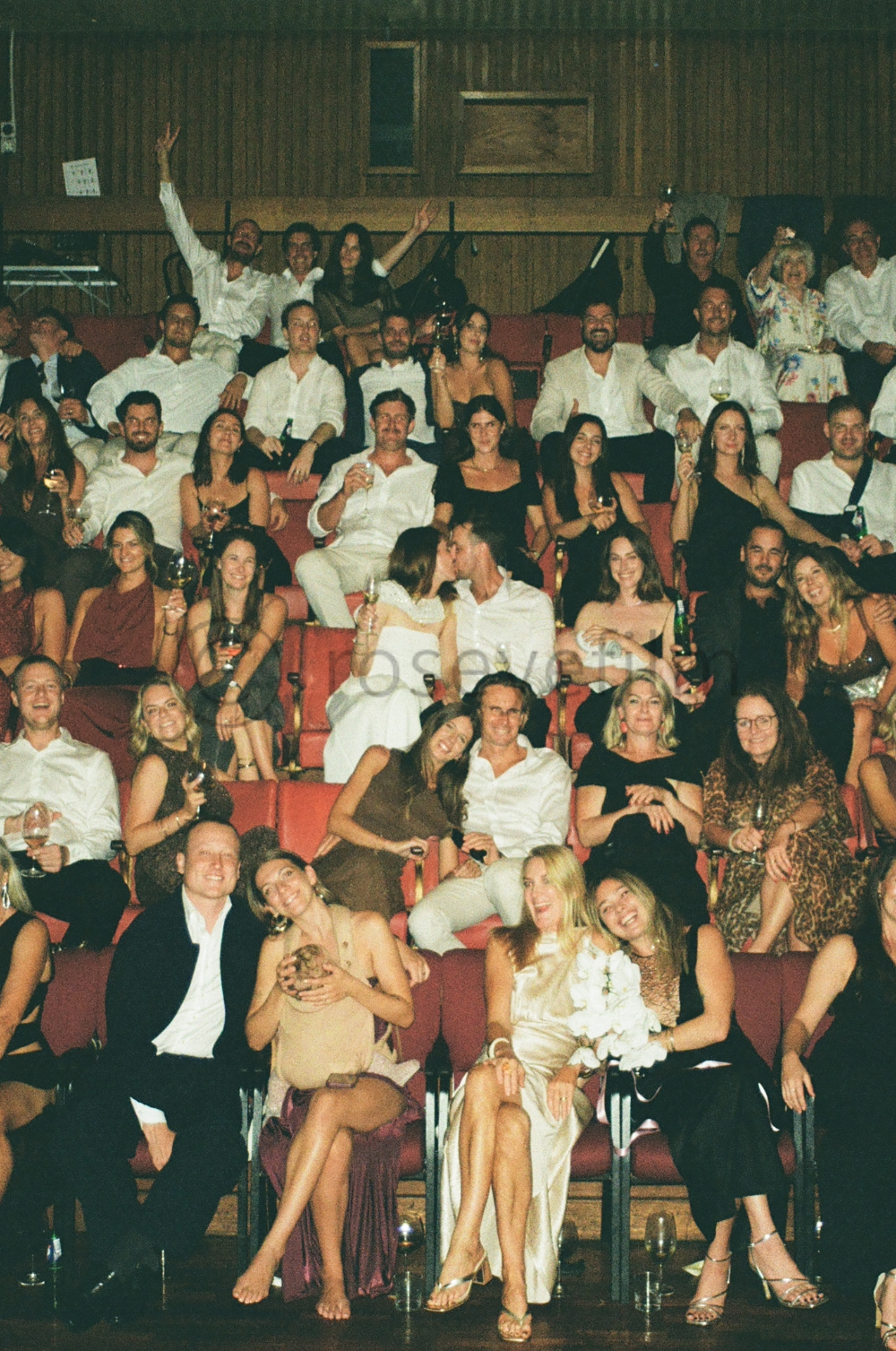 Group of people at a formal event sitting in auditorium seats, dressed in elegant attire, holding drinks, smiling and making celebratory gestures.