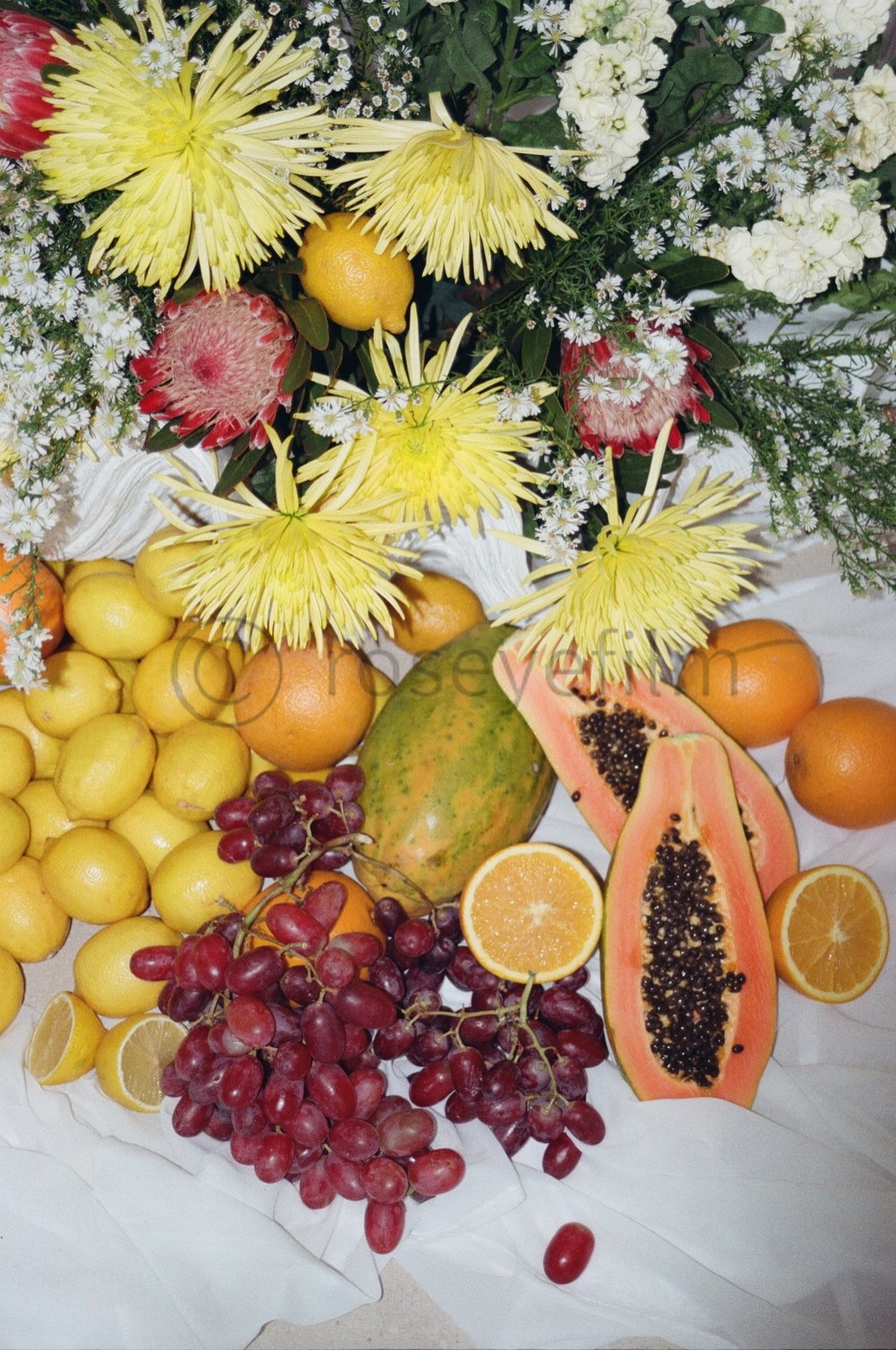 A colorful arrangement of various fresh fruits including grapes, lemons, oranges, a papaya, a mango, and a halved grapefruit, topped with a large bouquet of white, yellow, and pink flowers.
