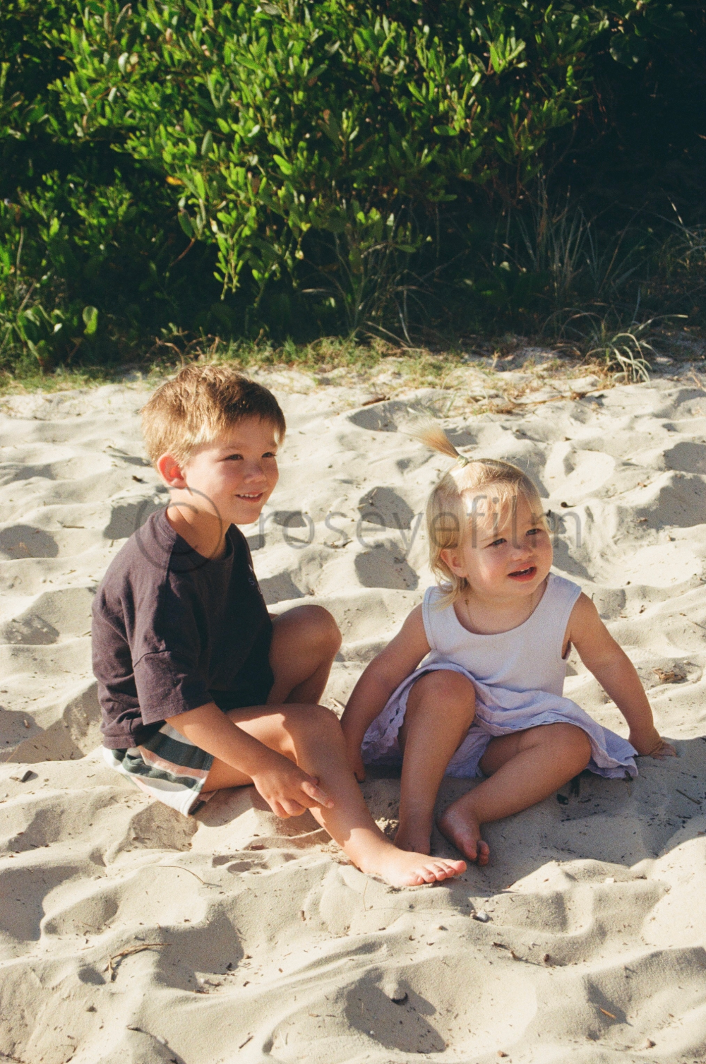 Two young children, a boy and a girl, sitting on sandy beach near green bushes. The boy is wearing a dark t-shirt and shorts, smiling while looking to the side. The girl is wearing a white dress, looking slightly distressed, with her hair tied up in 