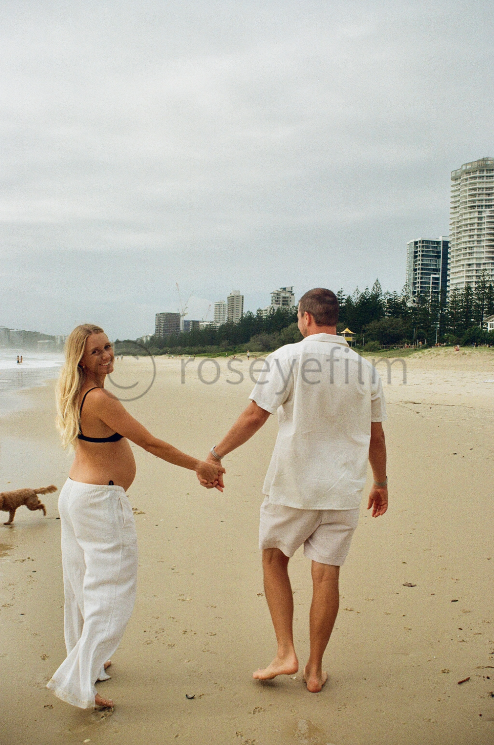 A couple holding hands on the beach, with a woman in a black bikini top and white pants and a man in a white shirt and beige shorts, with a dog in the background and city buildings in the distance under a cloudy sky.