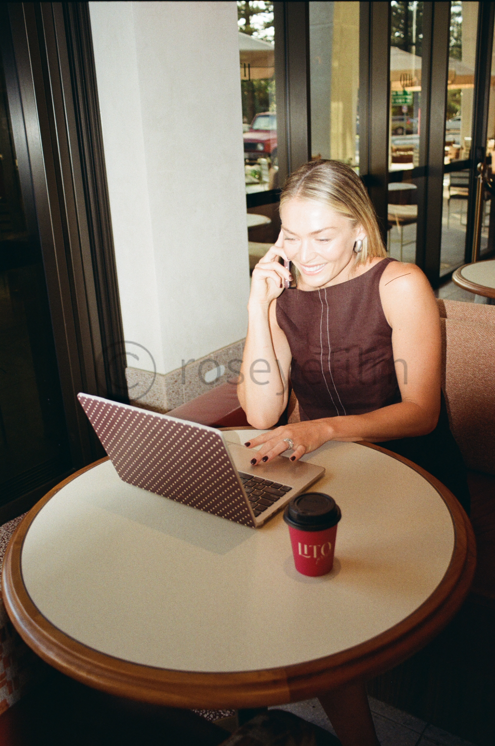 A woman with blonde hair sitting at a round café table, talking on her cellphone while using her laptop, with a coffee cup in front of her, in an indoor café with large windows.