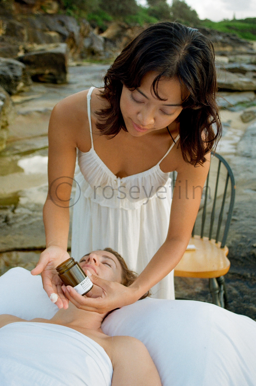 A woman giving a massage by a river, applying cream from a jar on a woman lying on a massage table, with rocks and greenery in the background.