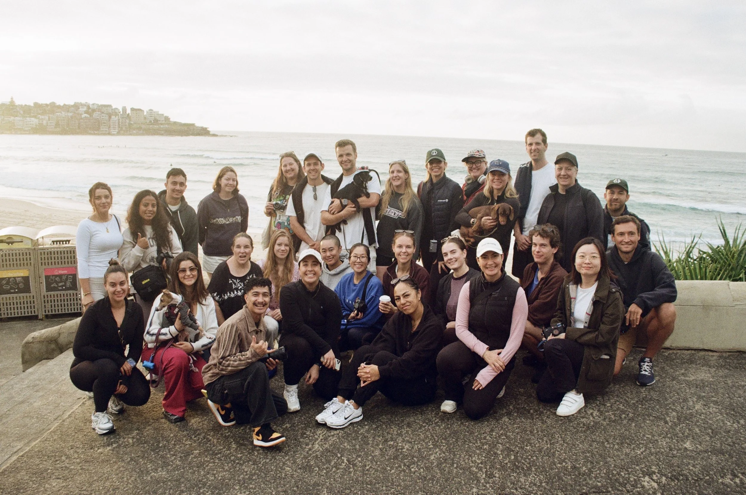 Group of people posing on a beach with the ocean in the background. They are smiling and holding cameras and dogs.