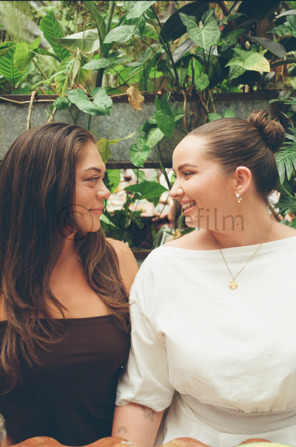 Two women smiling at each other, standing close together in front of green plants.