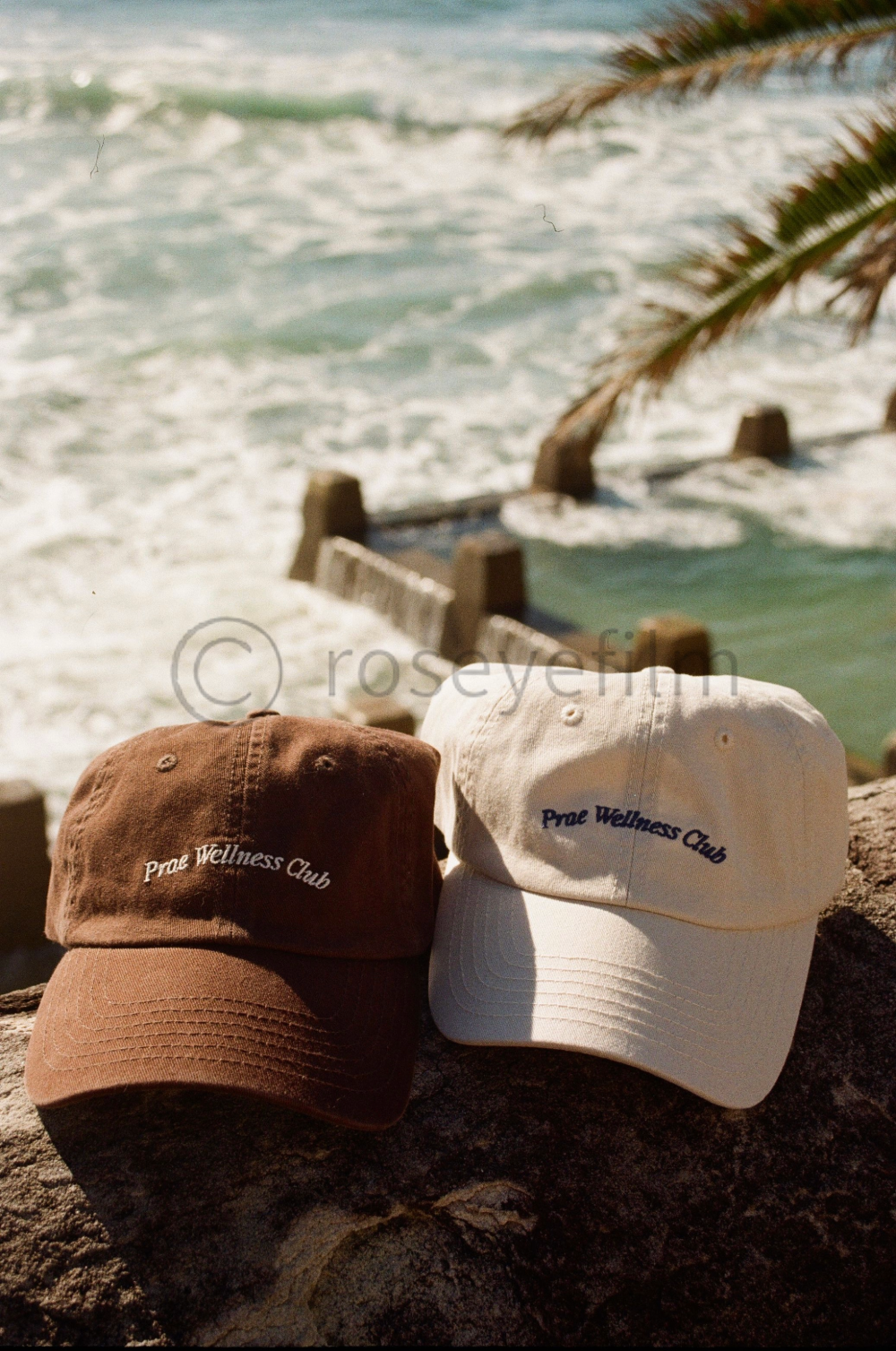 Two baseball caps, one brown and one beige, with 'Prae Wellness Club' embroidered on each, placed on a rock at a beachside location with ocean waves, a wooden breakwater, and palm leaves in the background.