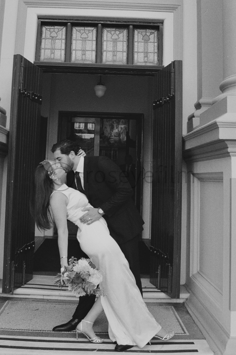 A man and woman sharing a dance inside a building with open doors, with the woman holding a bouquet of flowers and both leaning in for a kiss.
