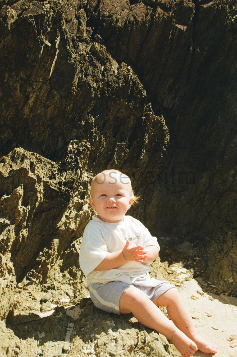 Young child sitting on sandy ground near large rock formation, smiling and looking upwards, dressed in a white t-shirt and gray shorts, with sunlight illuminating the scene.