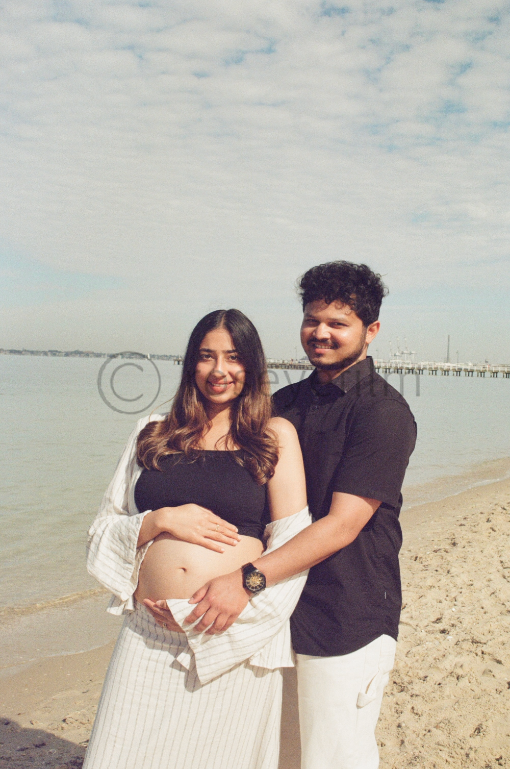 A pregnant woman and a man standing on a beach, smiling, with the woman resting her hand on her belly and the man holding her waist, under a cloudy sky.