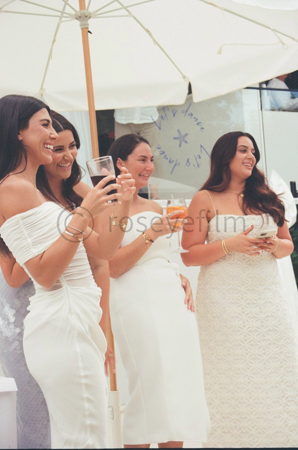 Four women in white dresses smiling and holding drinks at an outdoor event, under a white umbrella.