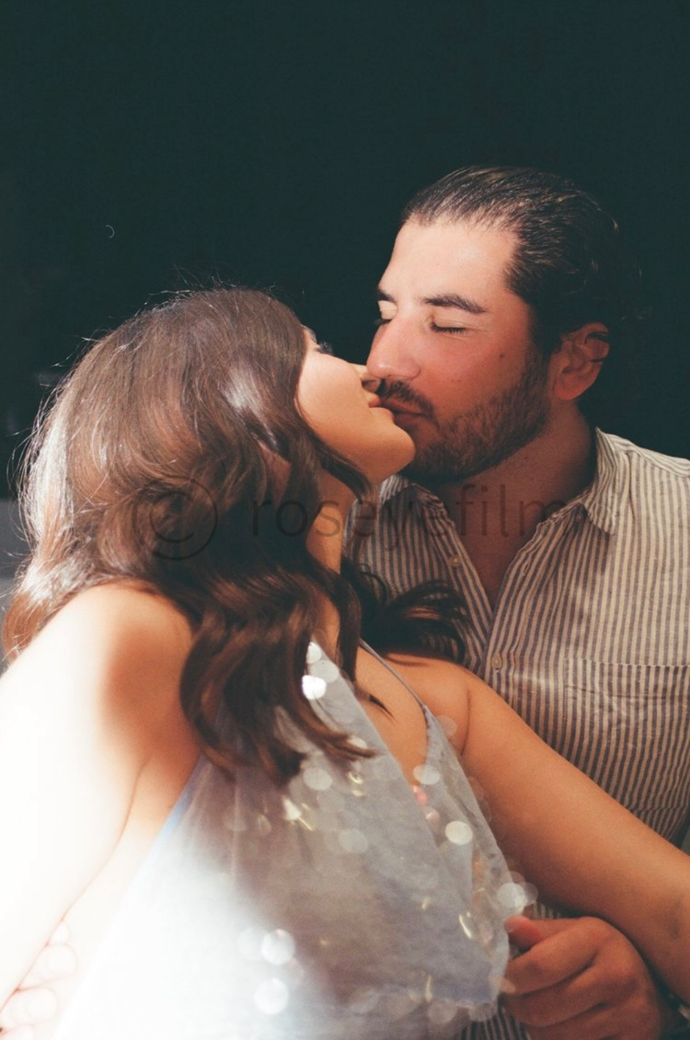 A woman and a man kissing each other, with their eyes closed, in a romantic setting with soft lighting and a dark background.