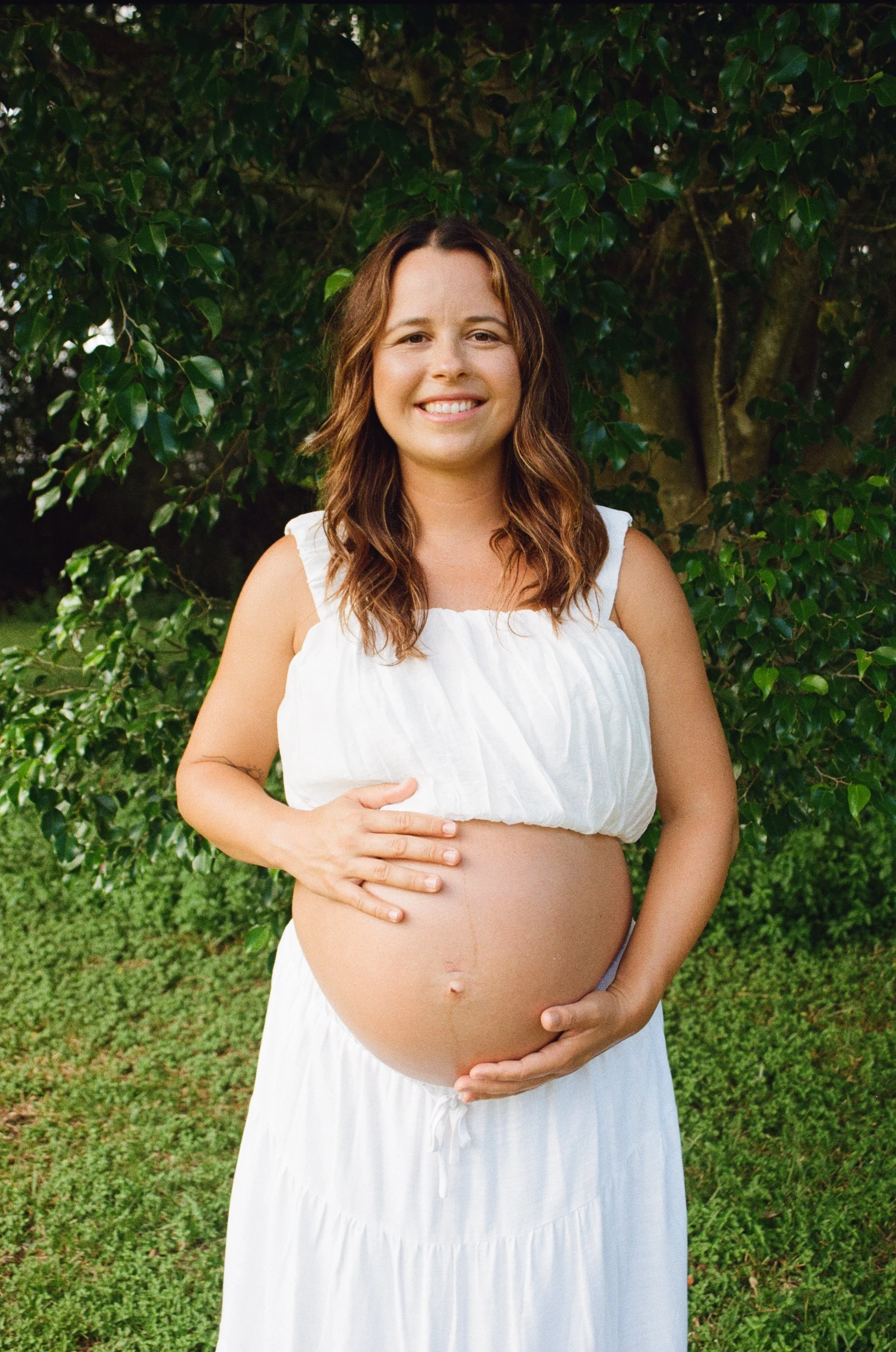 A pregnant woman with long brown hair wearing a sleeveless white dress, standing outdoors in front of green leafy bushes, smiling and holding her bare pregnant belly with both hands.
