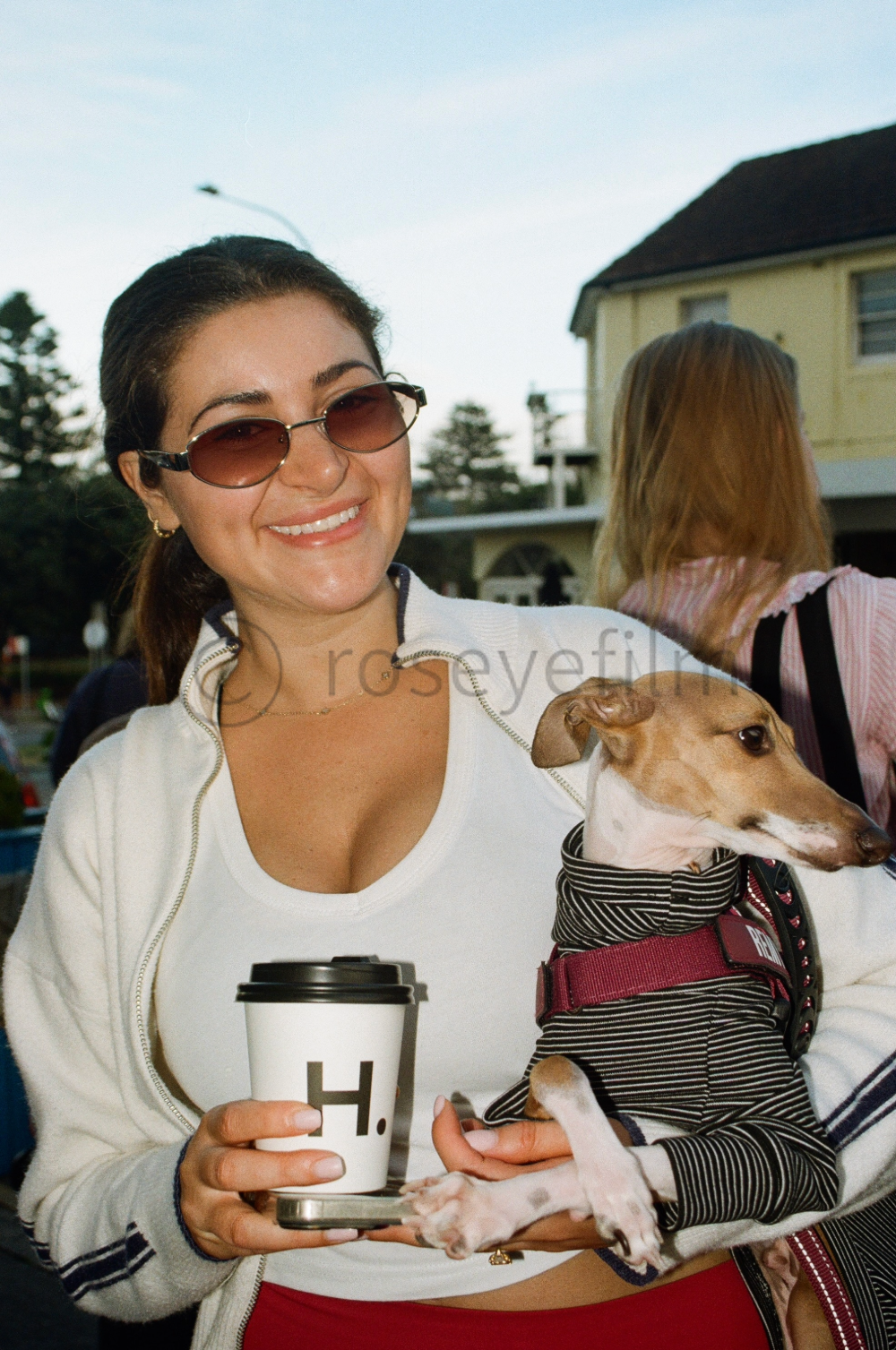 Smiling woman wearing sunglasses holding a small dog in a striped hoodie outdoors.