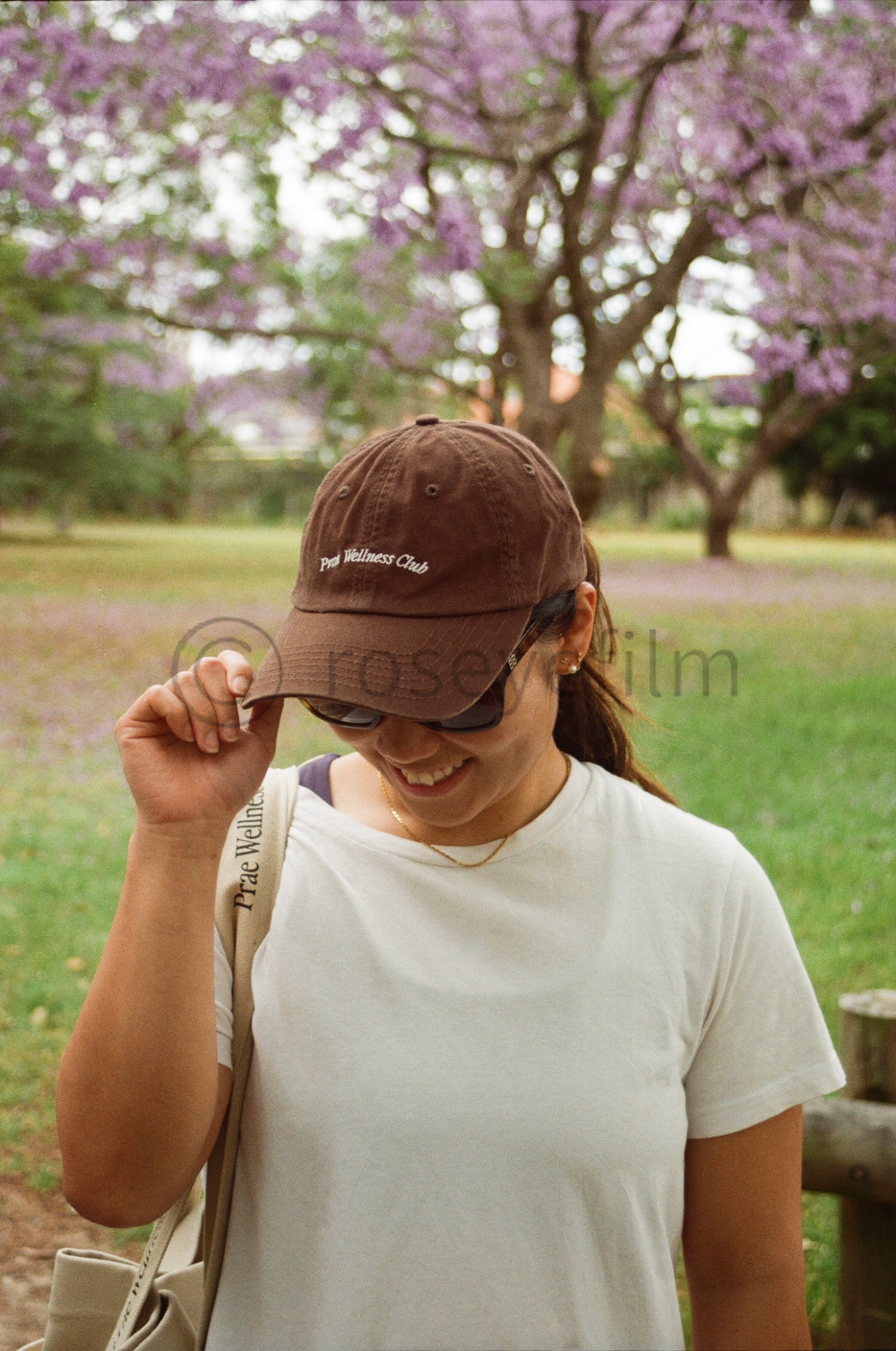 A woman wearing a brown cap with white text reading "Prae Wellness Club," sunglasses, and a white t-shirt is smiling and adjusting her cap in a park with blooming pink and purple trees in the background.