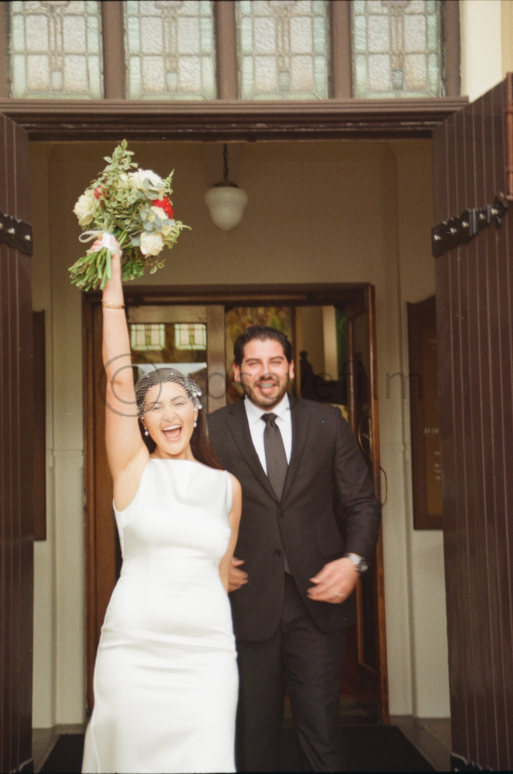A bride and groom celebrating as they walk out of a building, with the bride holding a bouquet of flowers in the air and smiling joyfully.