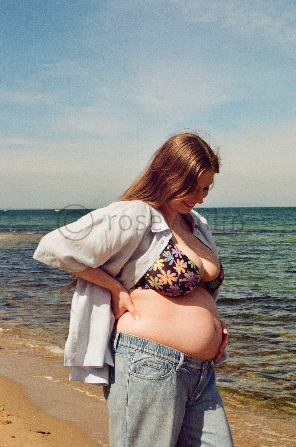 A pregnant woman with long brown hair standing on a beach, wearing a floral bikini top, loose jeans, and an unbuttoned shirt, looking down at her belly with a smile.