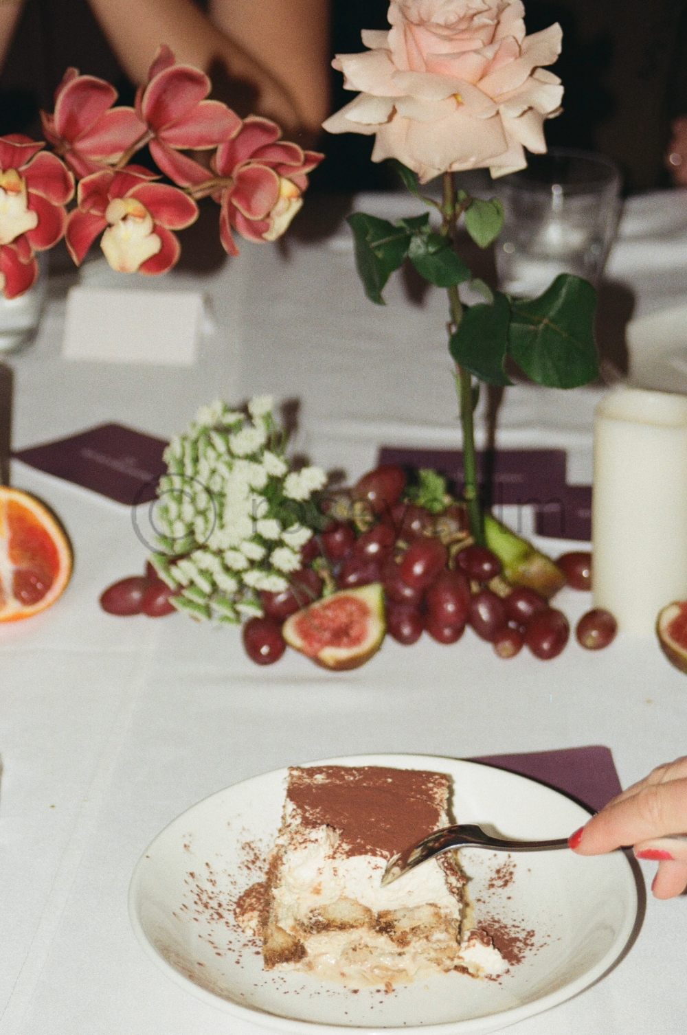 A plate with a partially eaten slice of tiramisu with cocoa powder, on a white table. There are pink and cream roses, red grapes, figs, and a glass candle holder on the table.